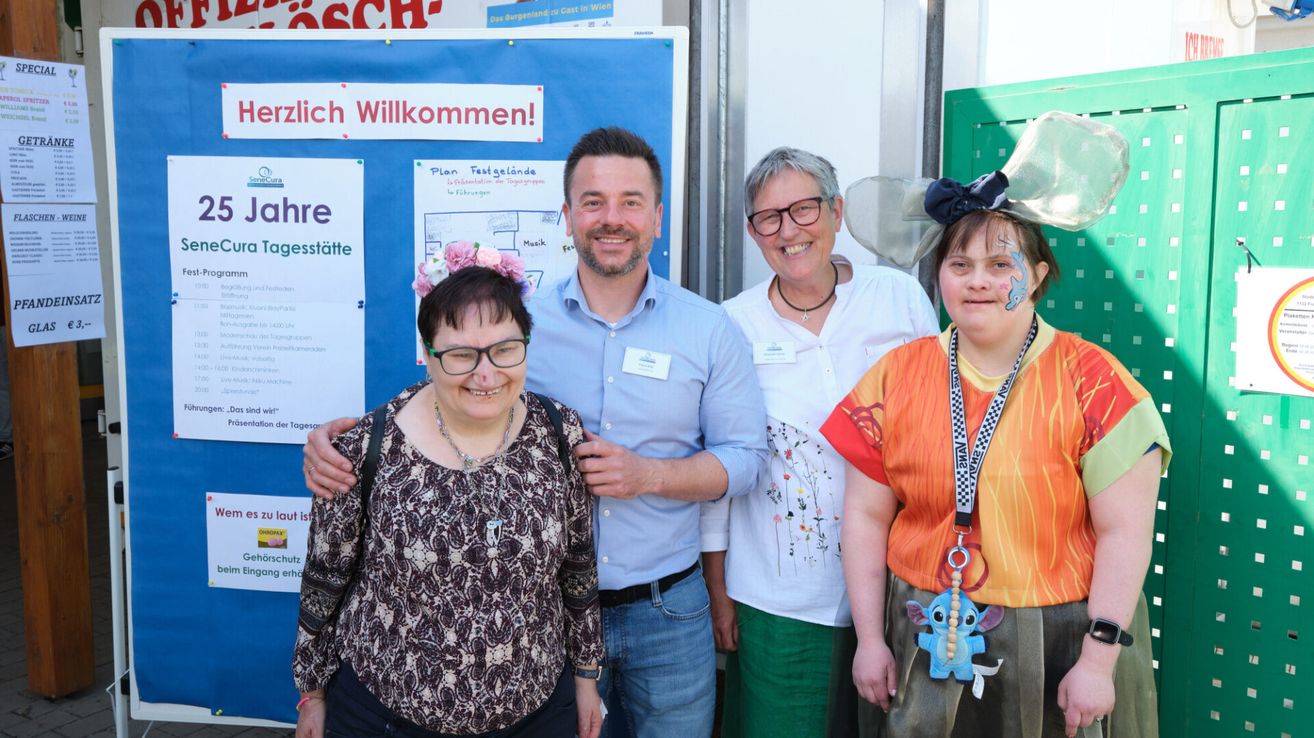 Eine Gruppe von Menschen steht vor einem blauen Banner mit Text. Die Frau links trägt eine Brille und eine Blumenkopfbedeckung. Der Mann in der Mitte lächelt und trägt ein blaues Hemd. Die Frau rechts trägt eine Halskette und hat Gesichtsbemalung. Die Frau hinter ihnen trägt eine Brille und ein weißes Hemd.