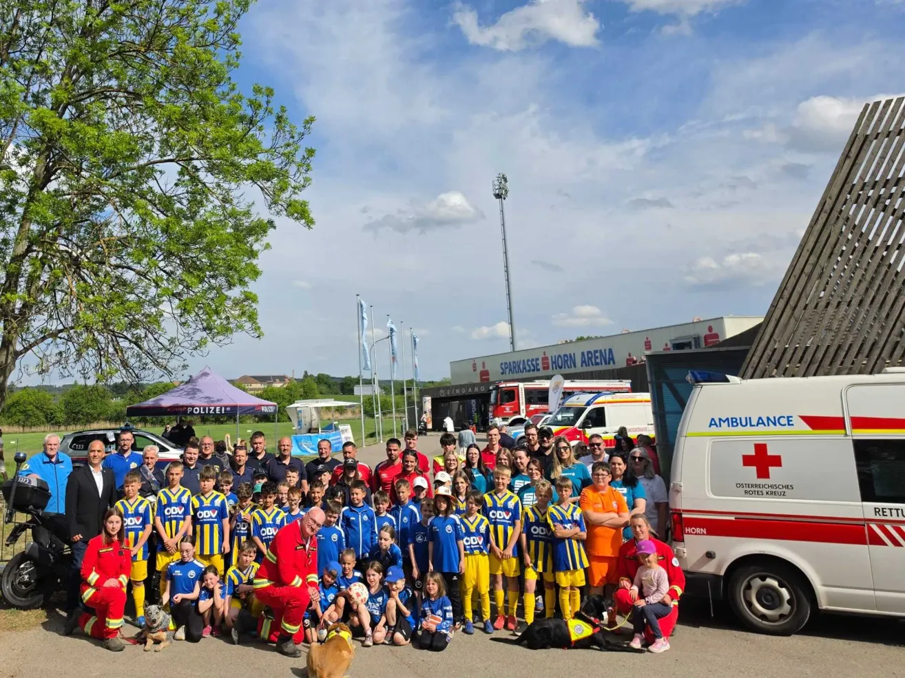 Eine Gruppe von Kindern, Erwachsenen und einem Feuerwehrmann posiert für ein Foto vor einem Krankenwagen und einem Gebäude. Einige tragen blaue und gelbe Uniformen, und ein Baum ist im Hintergrund zu sehen.