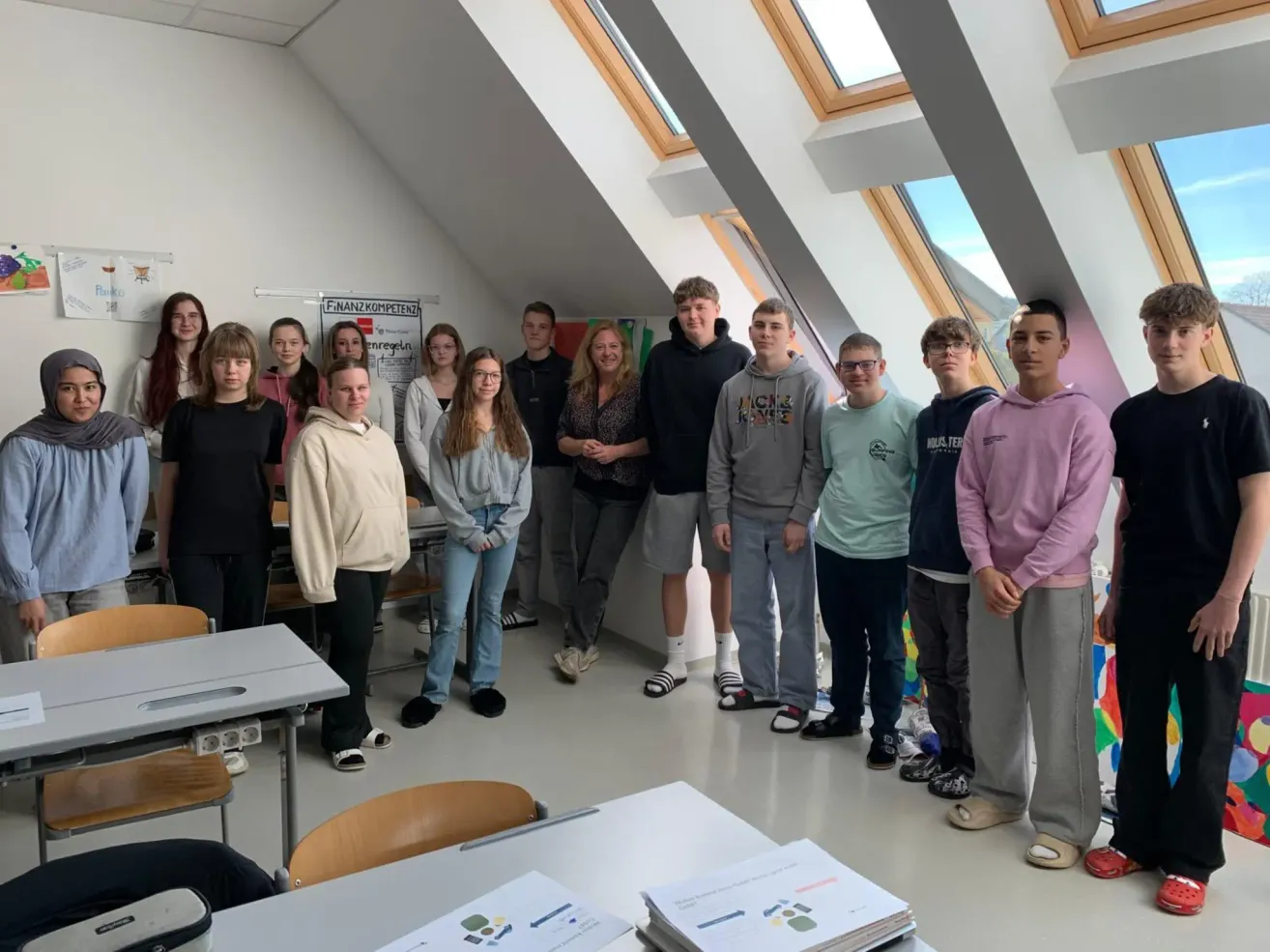 Eine Gruppe von Schülern und Lehrern posiert für ein Foto in einem Klassenzimmer. Es stehen zwei Erwachsene in der vorderen Reihe und einige Schüler dahinter. Das Klassenzimmer hat Tische und Stühle, und es gibt Fenster an der Wand.