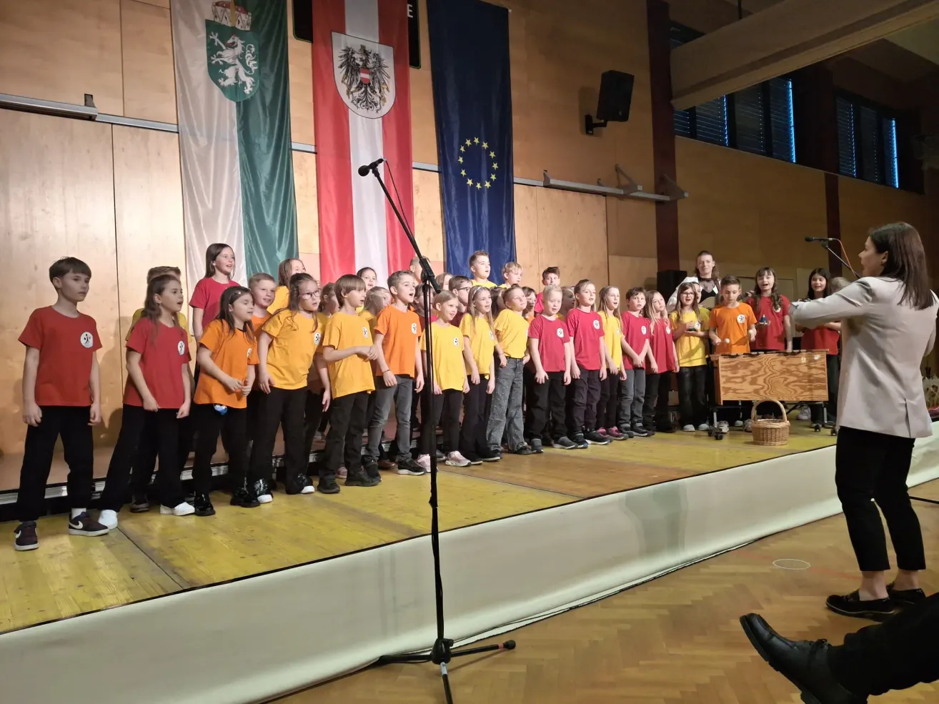 A group of young children in matching yellow and red shirts are singing on a stage with a microphone in front of them. Behind them are flags.