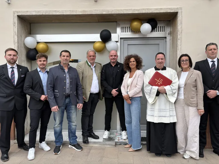 A group of individuals stands in front of a building. Among them, a priest holds a book, while others wear formal attire. Balloons are visible above the entrance.