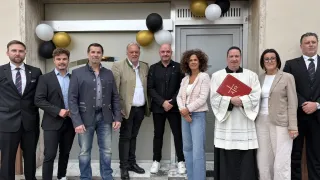 A group of individuals stands in front of a building. Among them, a priest holds a book, while others wear formal attire. Balloons are visible above the entrance.