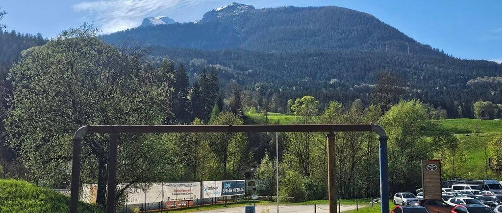 Ein landschaftlicher Blick auf einen Berg mit schneebedecktem Gipfel, umgeben von grünen Bäumen und Pflanzen. Es gibt ein blaues Banner mit Text und eine Metallstruktur im Vordergrund.