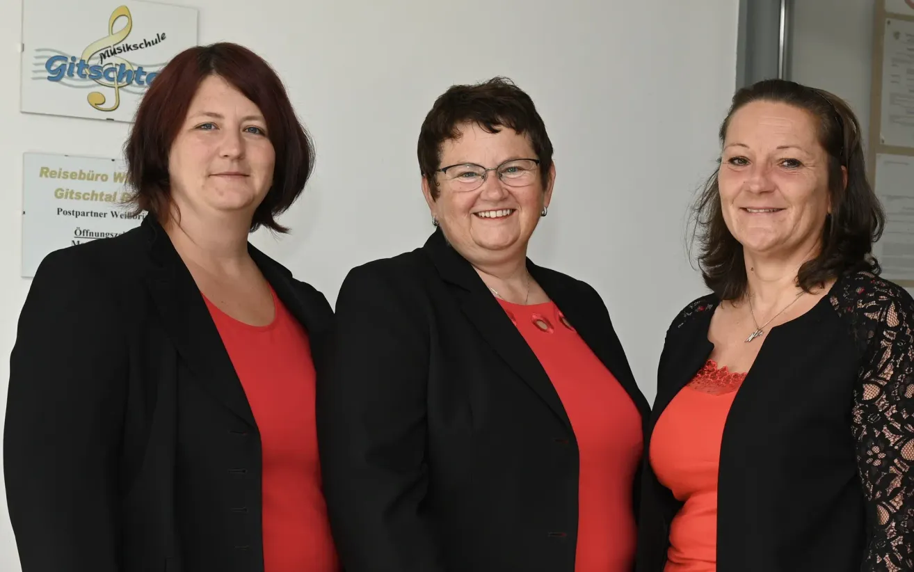 Drei Frauen stehen nebeneinander und lächeln in die Kamera. Die Frau in der Mitte trägt eine Brille und eine Halskette. Sie tragen alle rote Oberteile und schwarze Jacken.