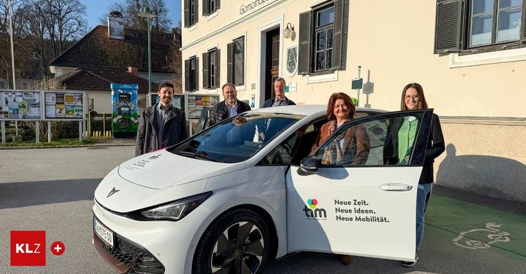 Fünf Personen posieren neben einem weißen Elektroauto vor einem Gebäude mit einem Schild, das 'Neue Zeit. Neue Ideen. Neue Mobilität.' besagt.
