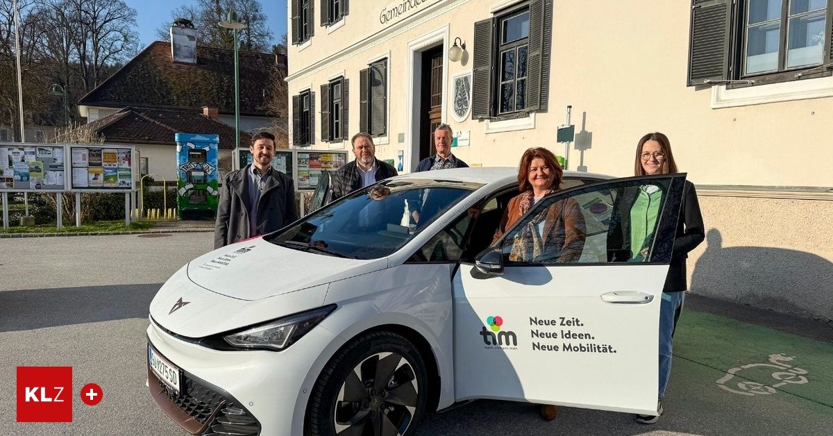 Fünf Personen posieren neben einem weißen Elektroauto vor einem Gebäude mit einem Schild, das 'Neue Zeit. Neue Ideen. Neue Mobilität.' besagt.