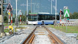A MAN electric bus drives on a railway track, with mountains and green trees in the background.