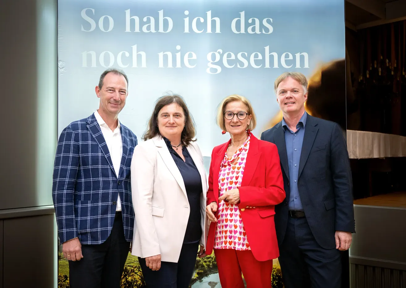 Four people stand before a sign with the words So hab ich das noch nie gesehen in white letters. They are all smiling. The woman in the center wears a red suit.
