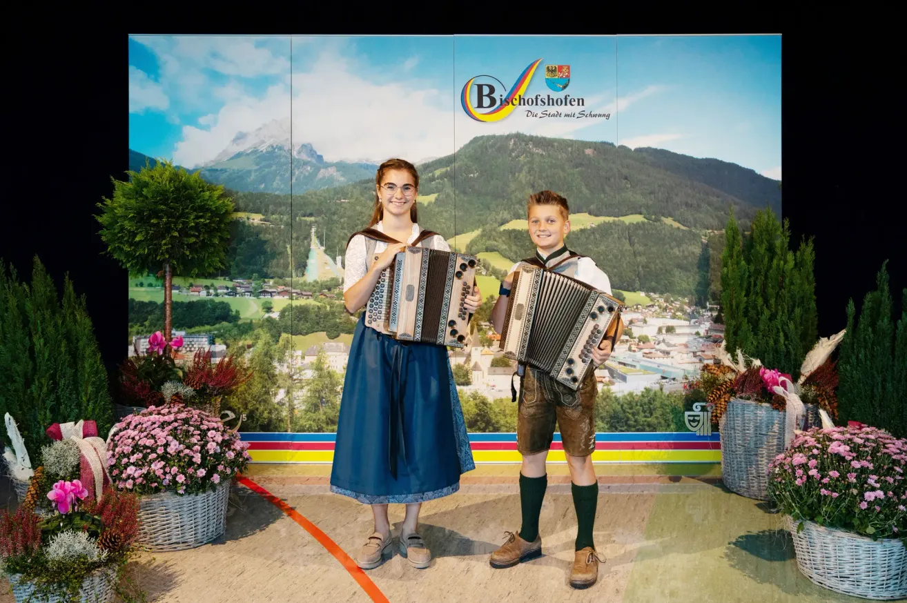 Zwei Kinder, ein Mädchen und ein Junge, stehen nebeneinander und halten ein Akkordeon. Sie sind in traditioneller bayerischer Kleidung gekleidet, mit Bergen und einem malerischen Dorf im Hintergrund.