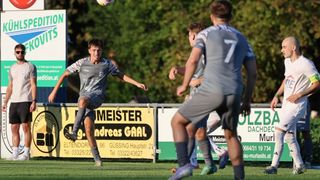 Drei Fußballspieler befinden sich auf einem Spielfeld, einer kickt den Ball. Der Spieler auf der linken Seite trägt ein Trikot mit der Nummer 7. Hinter ihnen befinden sich Banner mit Texten und Zahlen.
