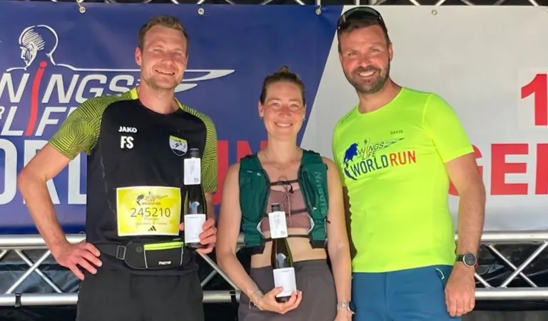 Three marathon runners pose for a photo with their awards; one holds a bottle of wine, another holds a medal. The runner in the middle wears a green vest.