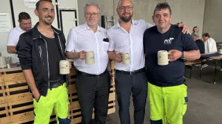 Four men pose for a picture, holding cups, two in reflective yellow safety gear, one in white with glasses, and another in blue with a logo on his shirt. They smile at the camera.