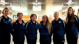 Four women in navy blue jackets with white stripes stand in a room. They are smiling and appear to be posing for a photo. Behind them is a yellow wall with a white sign.