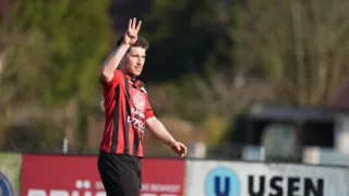 A soccer player in a red and black striped jersey makes a peace sign on a field. Trees and a banner are in the background.