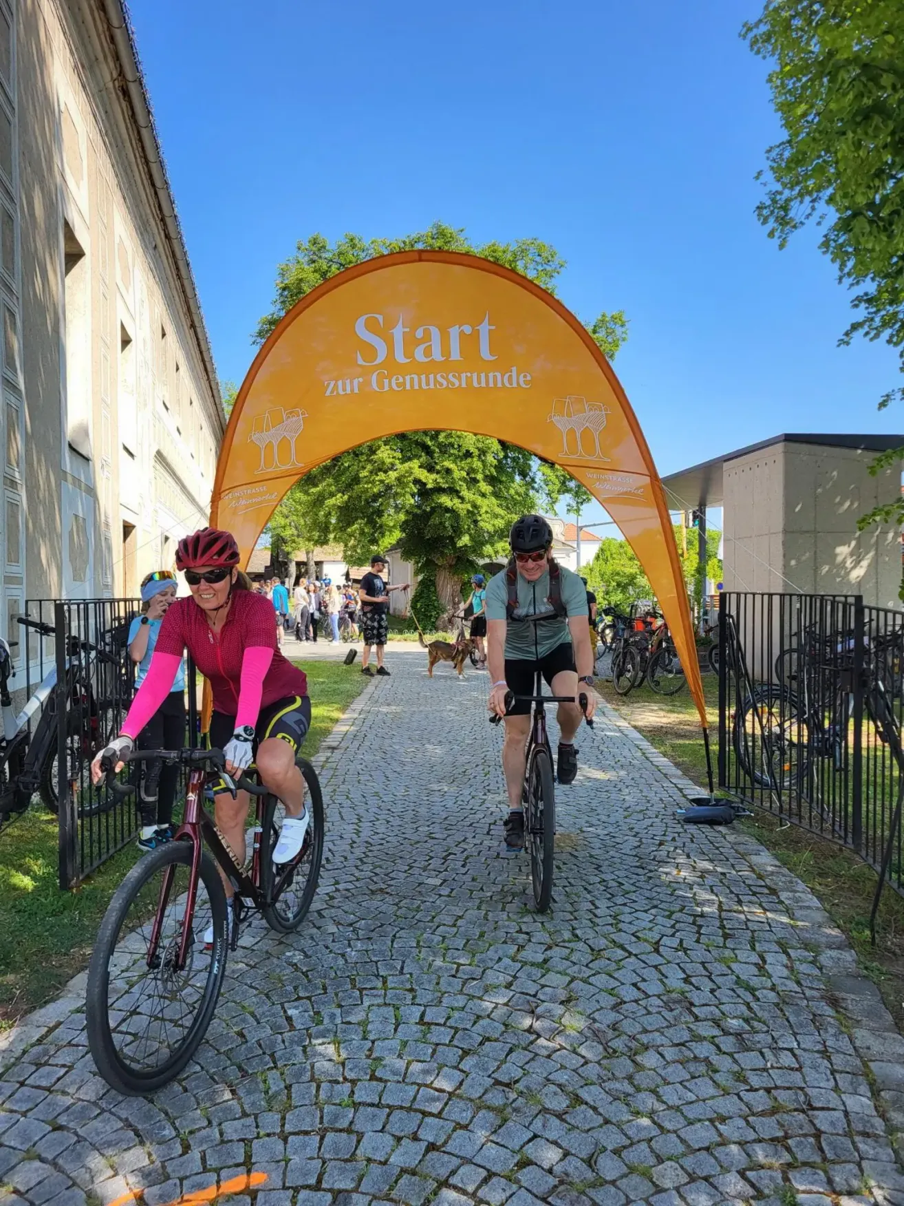 Zwei Radfahrer fahren unter einem gelben Banner, das Start zur Genussrunde steht. Sie sind auf einem Kopfsteinpflasterweg in der Nähe eines Gebäudes mit einem vorbeigehenden Hund.