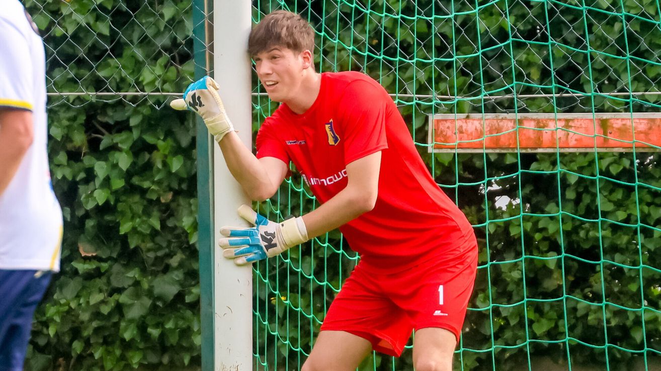Ein Fußballspieler in einem roten Trikot, das einen weißen Handschuh hält, steht bei einem Torpfosten. Die Nummer 1 ist auf seiner Shorts.