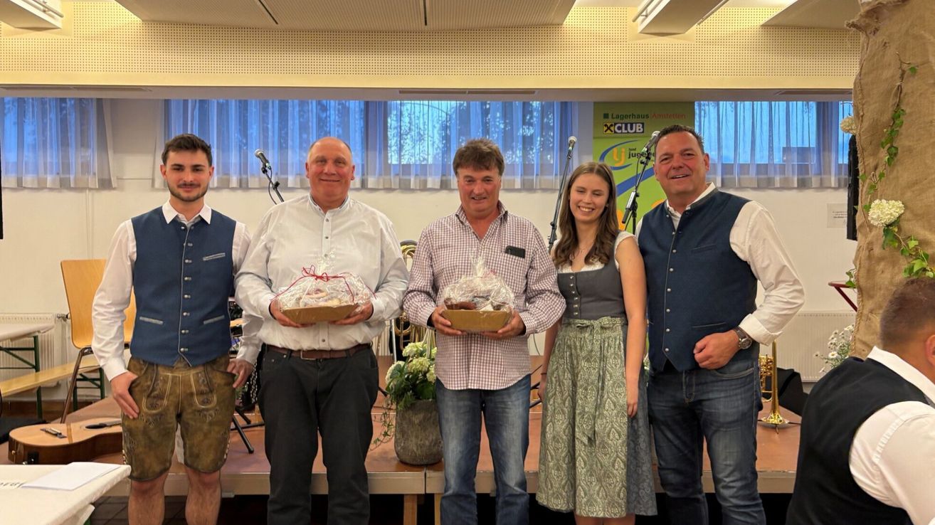 Five people pose for a photo, holding gifts. Two men wear casual shirts, one a checkered shirt. A woman in a dress stands between them. A sign with 'XCLUB' and 'Jung' is in the background.