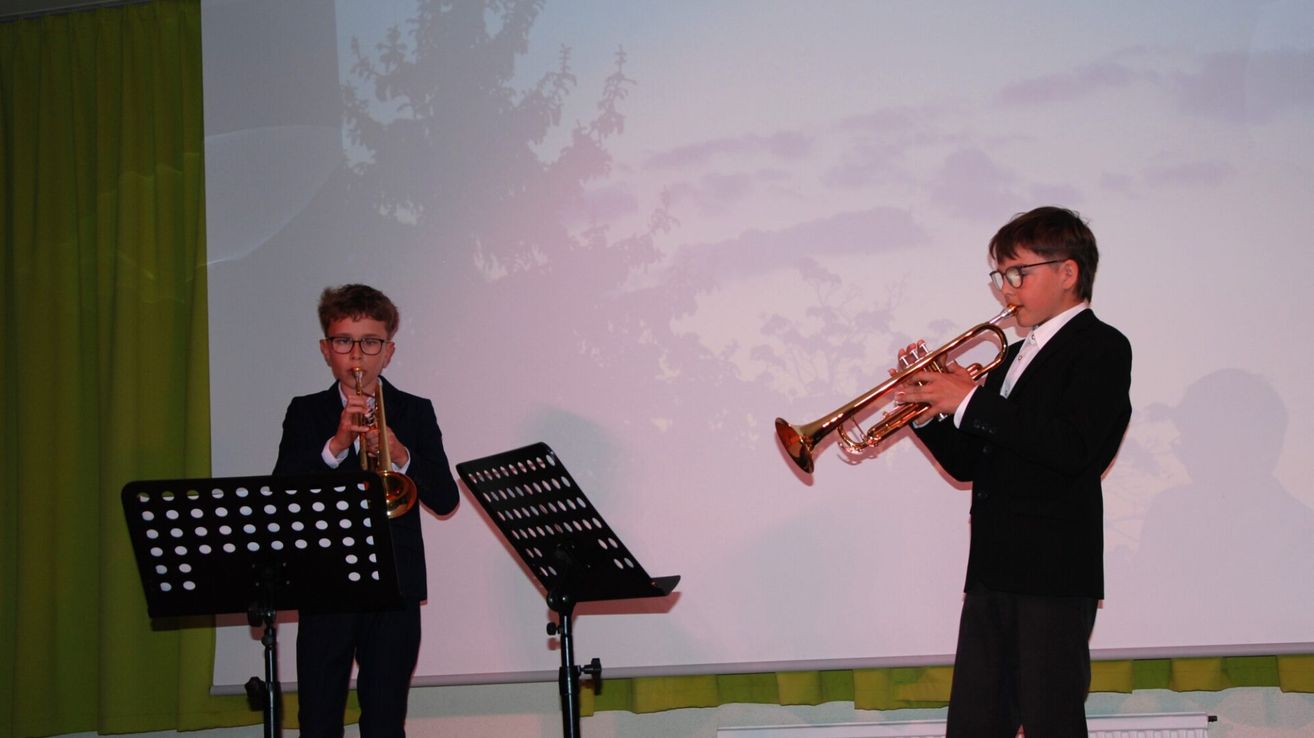 Two boys are playing trumpets on stage, one in front of a music stand and the other to the right. They are wearing formal attire. Behind them is a large screen displaying a pinkish sky with tree silhouettes.