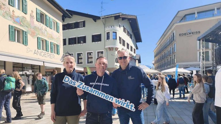 Drei Menschen in blauen Hoodies stehen an einem sonnigen Tag draußen und halten ein weißes Schild mit der Aufschrift 'Das Zusammen wirkt'. Hinter ihnen befinden sich Gebäude, darunter eines mit dem Schriftzug 'GERER'.