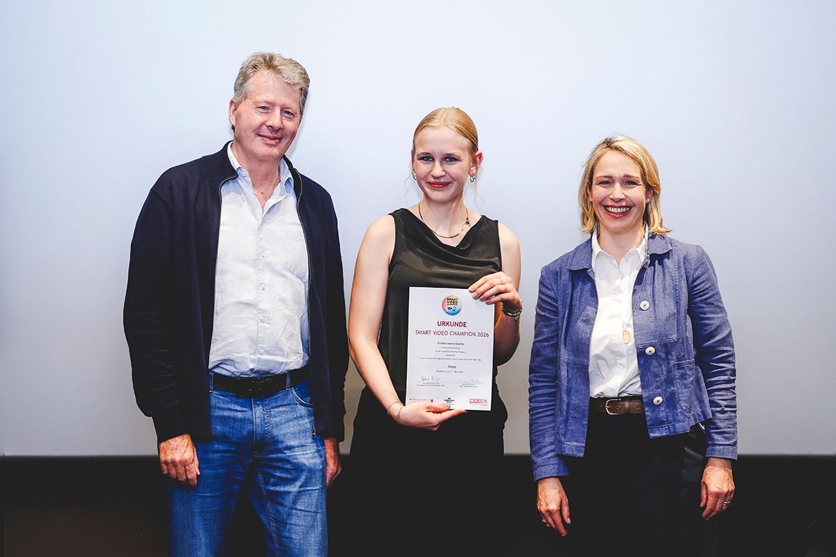 Three people stand together with a smiling woman holding a certificate. The man and woman on the side are smiling. They are in front of a white wall.