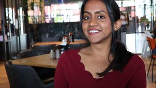 A woman with long black hair and a nose ring smiles at the camera in a restaurant. She wears a maroon top and a necklace with a diamond pendant. Behind her, tables and chairs are set up.