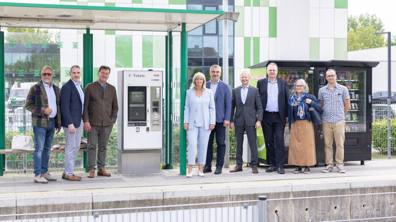 A group of people stands in front of a ticket vending machine at a bus stop, dressed formally. Behind them is a store with a green-and-white striped wall and a glass window.