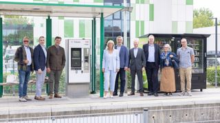 A group of people stands in front of a ticket vending machine at a bus stop, dressed formally. Behind them is a store with a green-and-white striped wall and a glass window.