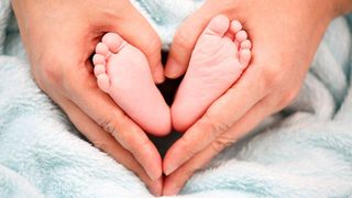 Two hands form a heart shape, holding a pair of newborn baby feet close together on a blue blanket.
