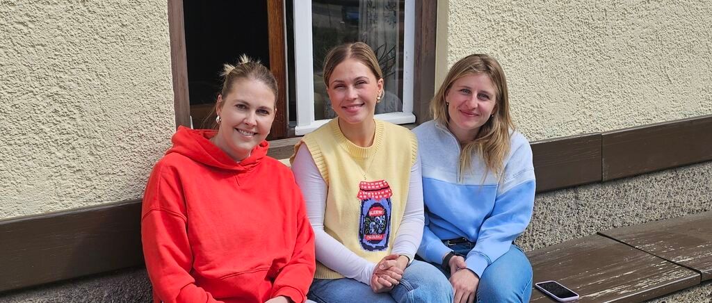 Drei Frauen sitzen auf einer Bank draußen. Eine trägt eine rote Kapuzenjacke, eine eine gelbe Weste und die andere ein blaues Hemd. Im Hintergrund ist ein Gebäude mit einem Fenster zu sehen.