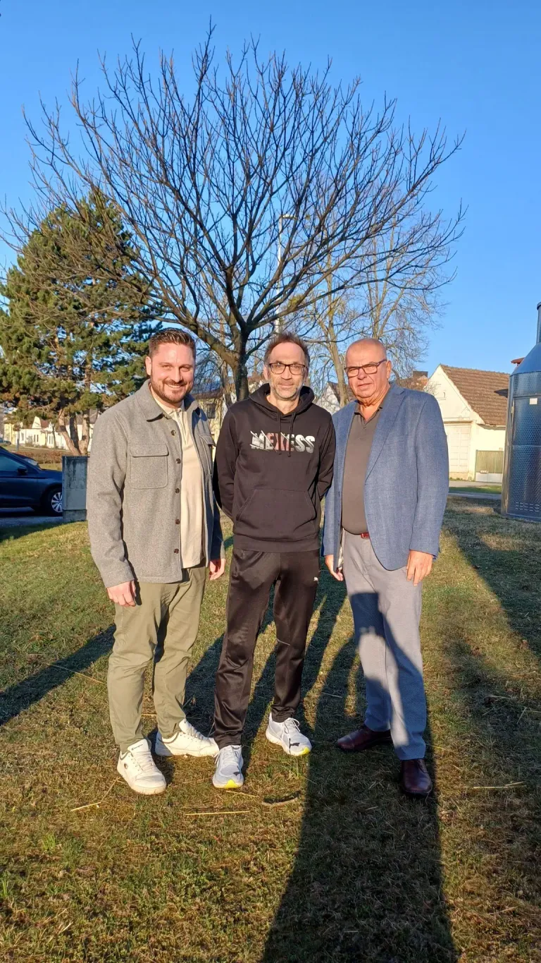 Three men stand outside on a grassy field. The man on the left smiles, while the middle man wears glasses and a hoodie. The man on the right wears a suit jacket. Behind them is a tree and houses.