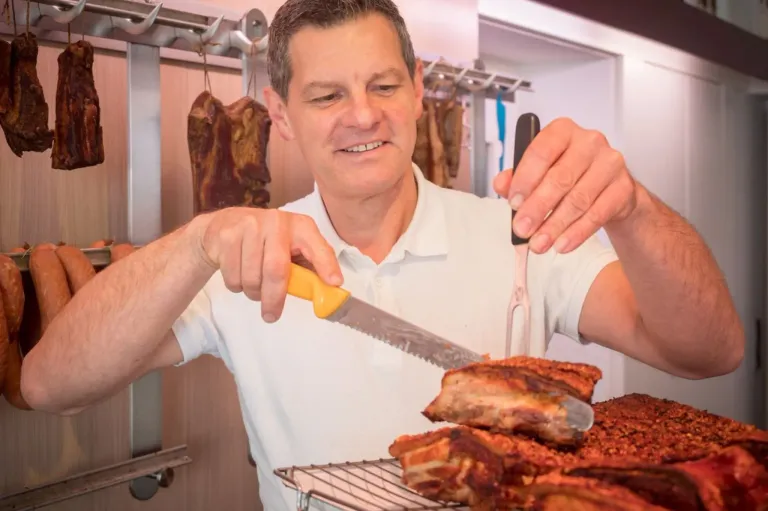 A man is cutting meat with a knife while holding a fork in his other hand. He is standing in front of a table with a rack of meat. There is a meat hanging from a hook.