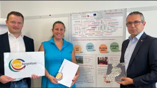 A woman in a blue shirt stands in front of a white poster. The poster has German text and includes various terms such as 'lebenswert,' 'heimat,' and 'blaufränkisch.' She holds two papers and smiles.