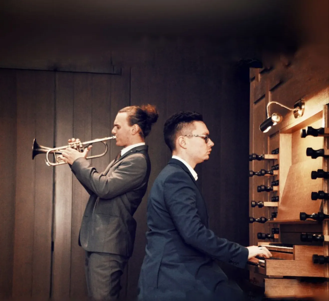 Zwei Männer in Anzügen spielen Musik, einer spielt Trompete und der andere an einer Orgel. Der Raum ist schwach beleuchtet.