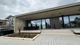 A modern building with large glass windows and a brown door. Outside, there are chairs and a small tree. The sky is cloudy.