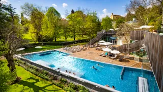 Ein Luftbild eines Resorts mit einem Außenpool, umgeben von einem üppigen grünen Rasen und Bäumen, und einem Gebäude mit Glaswänden.