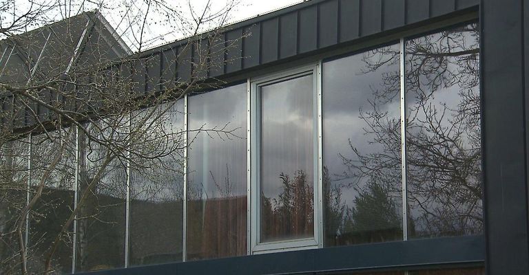 Ein modernes Gebäude mit grauer Außenseite und großen Glasfenstern. Die Fenster reflektieren nahe Bäume und einen bewölkten Himmel.