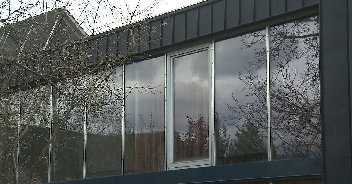 Ein modernes Gebäude mit grauer Außenseite und großen Glasfenstern. Die Fenster reflektieren nahe Bäume und einen bewölkten Himmel.