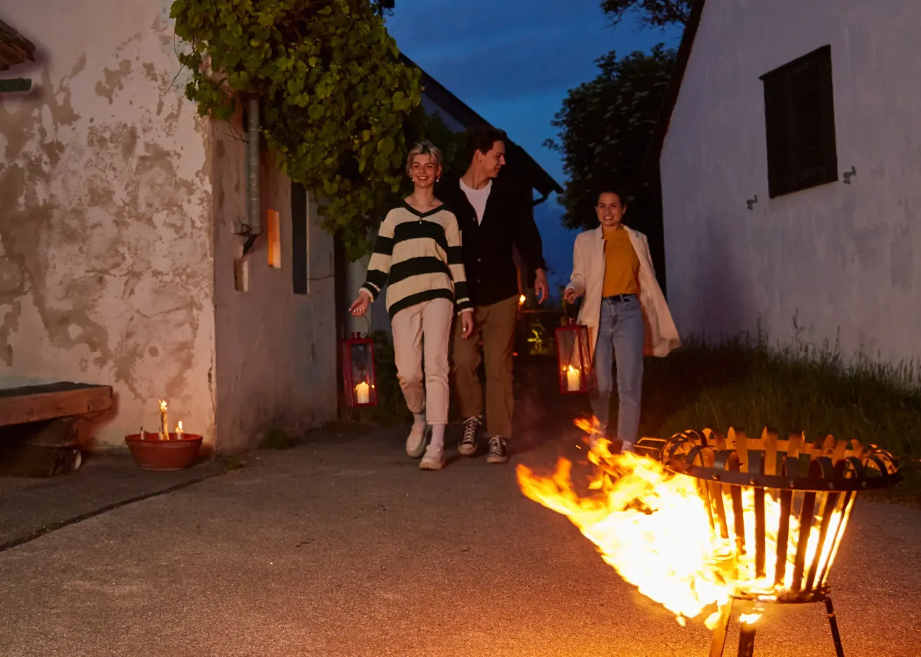 Drei Menschen gehen bei Nacht mit Laternen in der Hand auf ein Lagerfeuer zu. Sie lächeln und scheinen den Abend zu genießen.