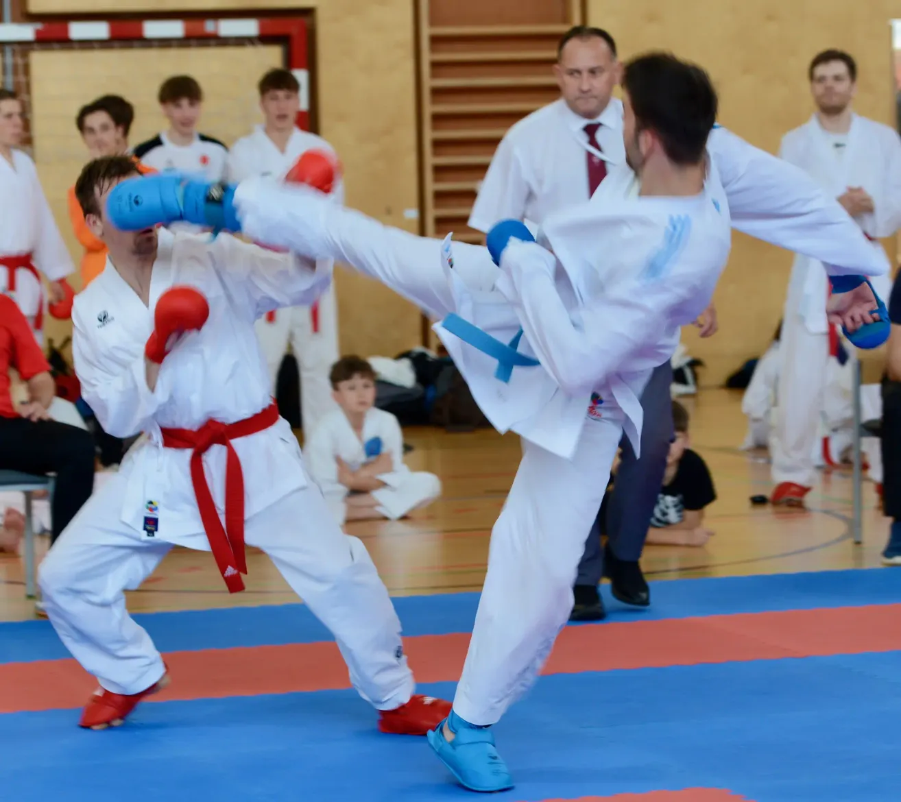 Two men are in a karate match, one wearing blue gloves and the other in red, both in white uniforms. Spectators watch from behind, some in white coats. The background features wooden panels and a blue-orange floor.