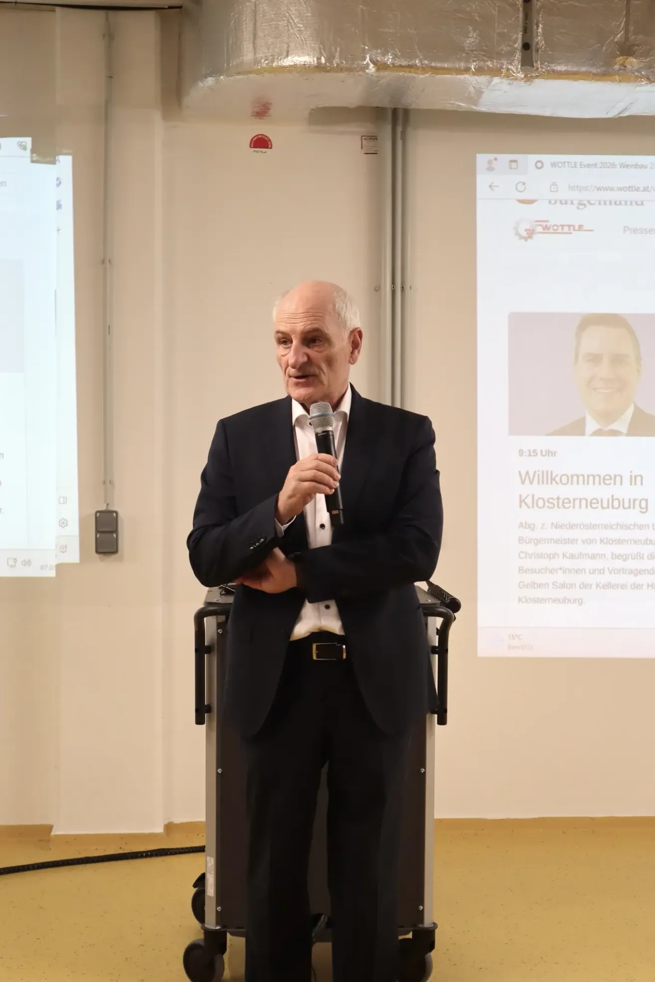 Ein Mann im Anzug steht mit einem Mikrofon am Rednerpult, vermutlich eine Rede haltend. Eine Projektionsleinwand hinter ihm zeigt ein Foto und Text.
