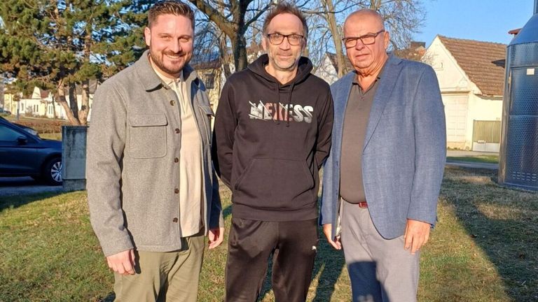 Three men stand in a park. The man on the left wears a gray jacket. The middle man wears a black hoodie with the word 'MEXISS' on it. The man on the right wears a blue blazer.