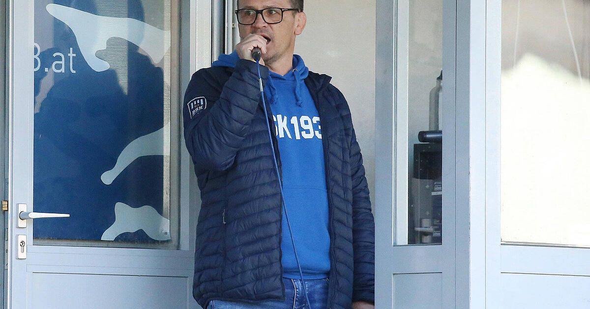 Ein Mann spricht in ein Mikrofon und trägt eine blaue Kapuzenjacke mit 'K1933' und einer dunklen Jacke. Die Szene spielt drinnen, möglicherweise bei einer Sportveranstaltung.