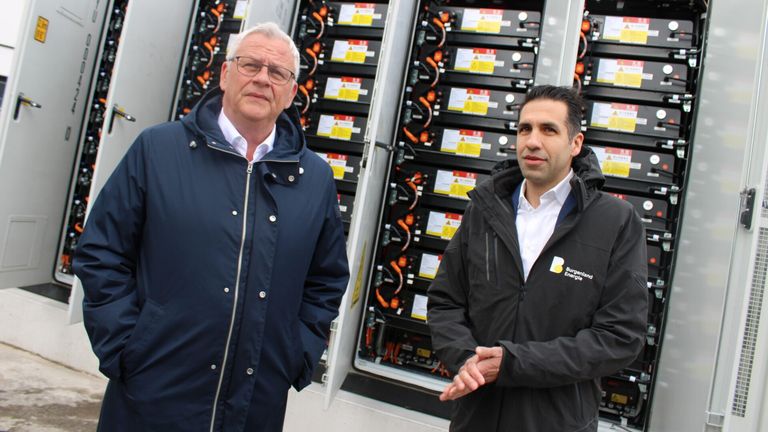 Zwei Männer stehen vor einem Batteriespeichersystem. Einer ist in einem blauen Mantel und der andere in einem schwarzen Jackett.