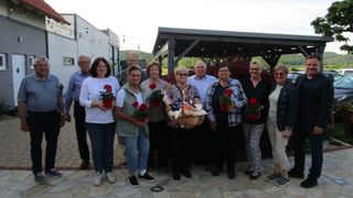 Eine Gruppe älterer Menschen steht zusammen, hält Blumen und einen Korb und posiert an einem sonnigen Tag für ein Foto.