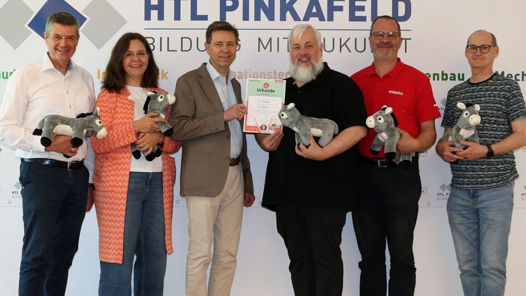 Vier Personen, die Stofftier-Esel halten, stehen gemeinsam für ein Foto. Der Mann in der Mitte hält ein Zertifikat und lächelt. Hinter ihnen steht ein Banner mit der Aufschrift 'HTL PINKAFELD BILDUNG MIT KUTUR.'