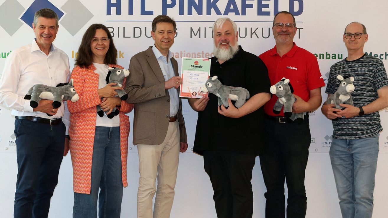Vier Personen, die Stofftier-Esel halten, stehen gemeinsam für ein Foto. Der Mann in der Mitte hält ein Zertifikat und lächelt. Hinter ihnen steht ein Banner mit der Aufschrift 'HTL PINKAFELD BILDUNG MIT KUTUR.'