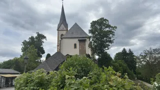 Eine kleine Kirche mit Turm und Uhr steht inmitten üppiger Bäume unter einem bewölkten Himmel.