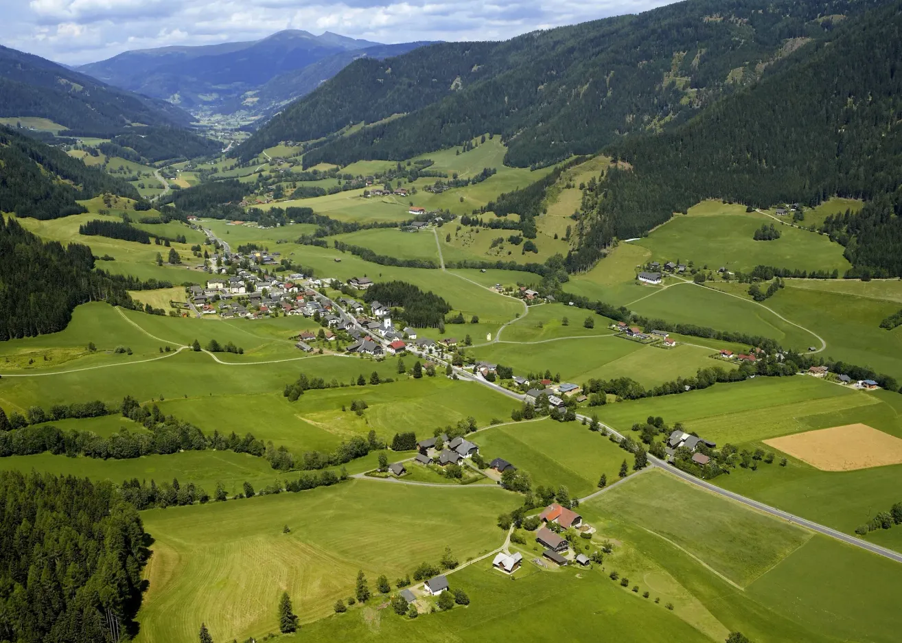Aerial view of a lush green valley with houses and roads nestled between hills and mountains under a cloudy sky.