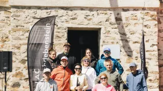 A group of people wearing hats and glasses are standing in front of a stone building with an open door. A speaker is on the left side.