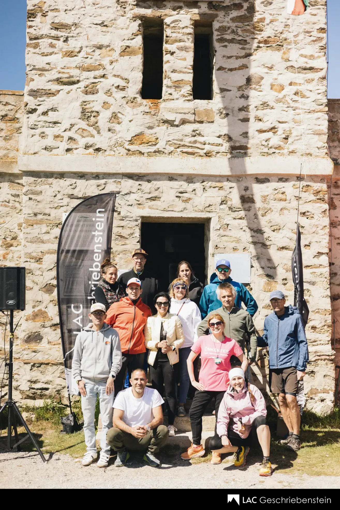 Eine Gruppe von Menschen mit Hüten und Brillen steht vor einem Gebäude aus Stein mit einer offenen Tür. Ein Lautsprecher befindet sich auf der linken Seite.